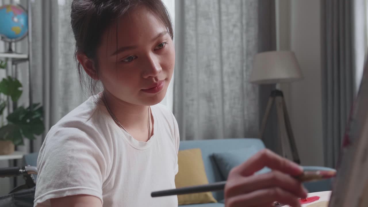 Asian Artist Girl In Wheelchair Holding Paintbrush And Mixing It With Colour Before Painting On The Canvas