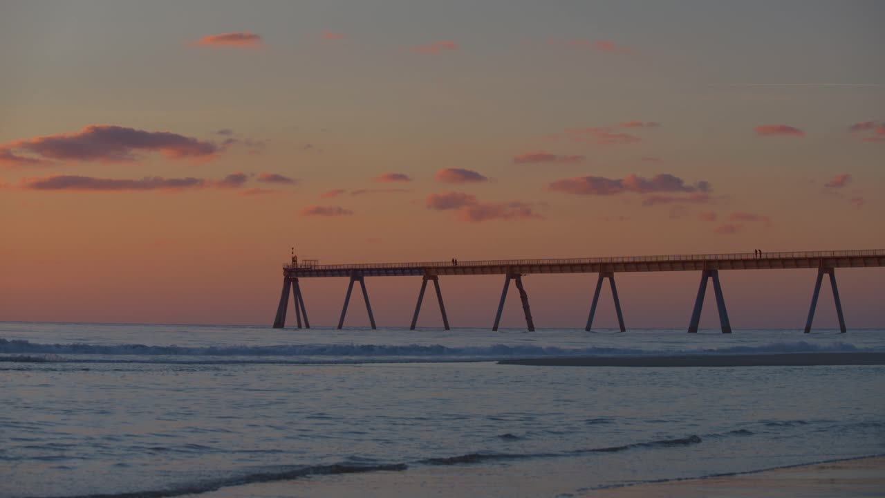 una puesta de sol naranja quemada en la playa con olas rodando y un largo muelle
