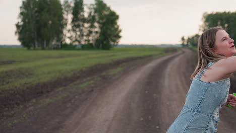 close up front view of woman in denim dress blowing soap bubbles from wand while smartphone peeks from dress pocket rustic dirt road farmland backdrop sunset light
