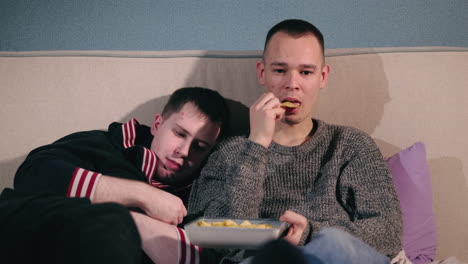 two men relaxing on a couch and eating chips