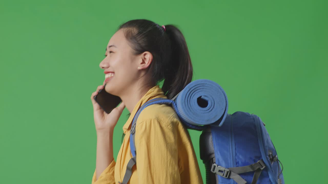 vista lateral de cerca de una excursionista asiática con mochila de montañismo hablando en teléfono inteligente y mirando a su alrededor mientras camina en el fondo de la pantalla verde en el estudio