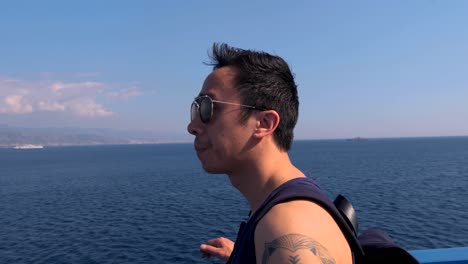 slow motion stablized circling shot of young vietnamese adult with aviator sunglasses soaking in view of sicily from ferry