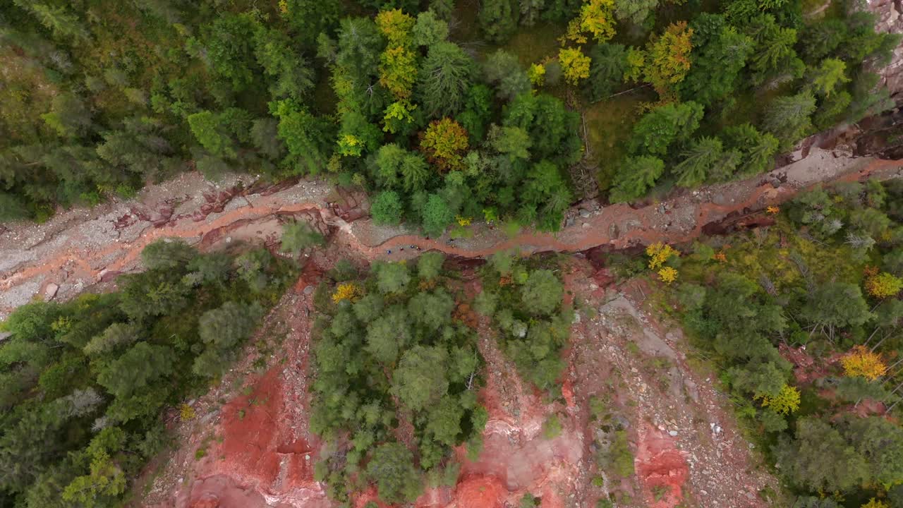 Top-down view of a red rock canyon with a group of hikers navigating the rugged terrain and jumping over a flowing creek in South Tyrol. Surrounded by dense forest and vibrant autumn colors.