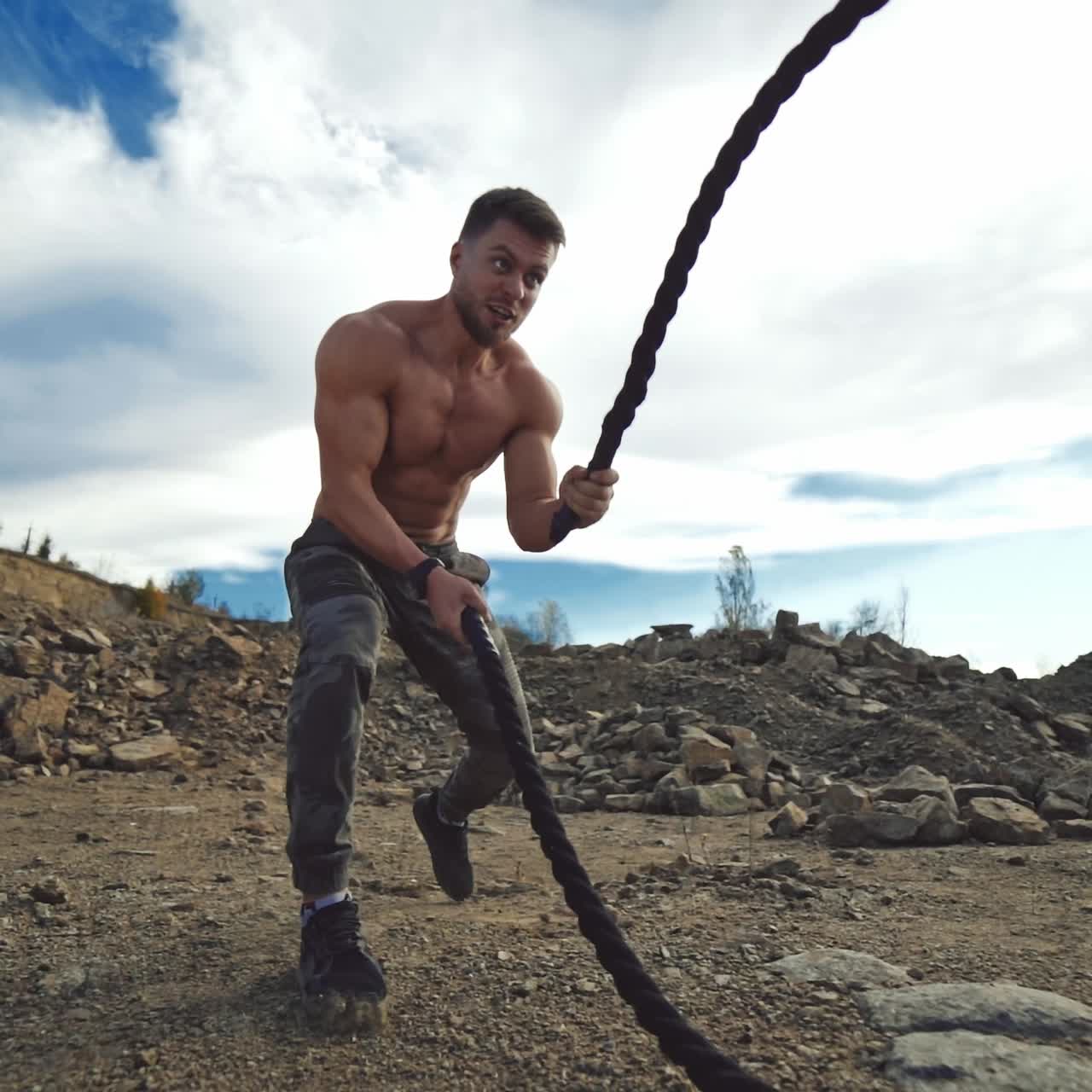 Cross power training. Concentrated sportsman exercises outdoors. Young shirtless muscular man training with battle ropes on rocky background. Slow Motion