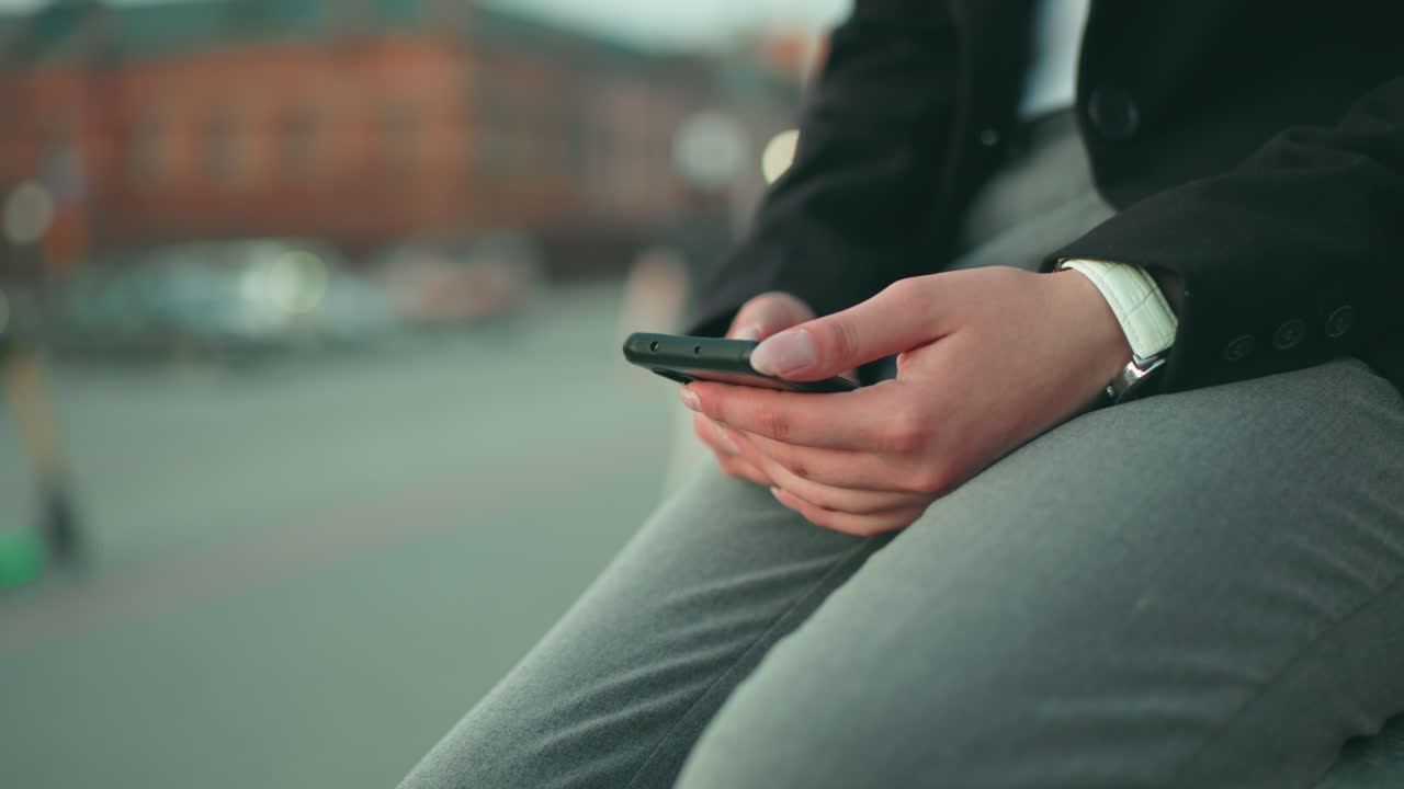 Close up partial view of lady in black suit seated with phone in hand, grey pants, and white watch, with blurred bokeh background showing people walking near buildings in busy city environment