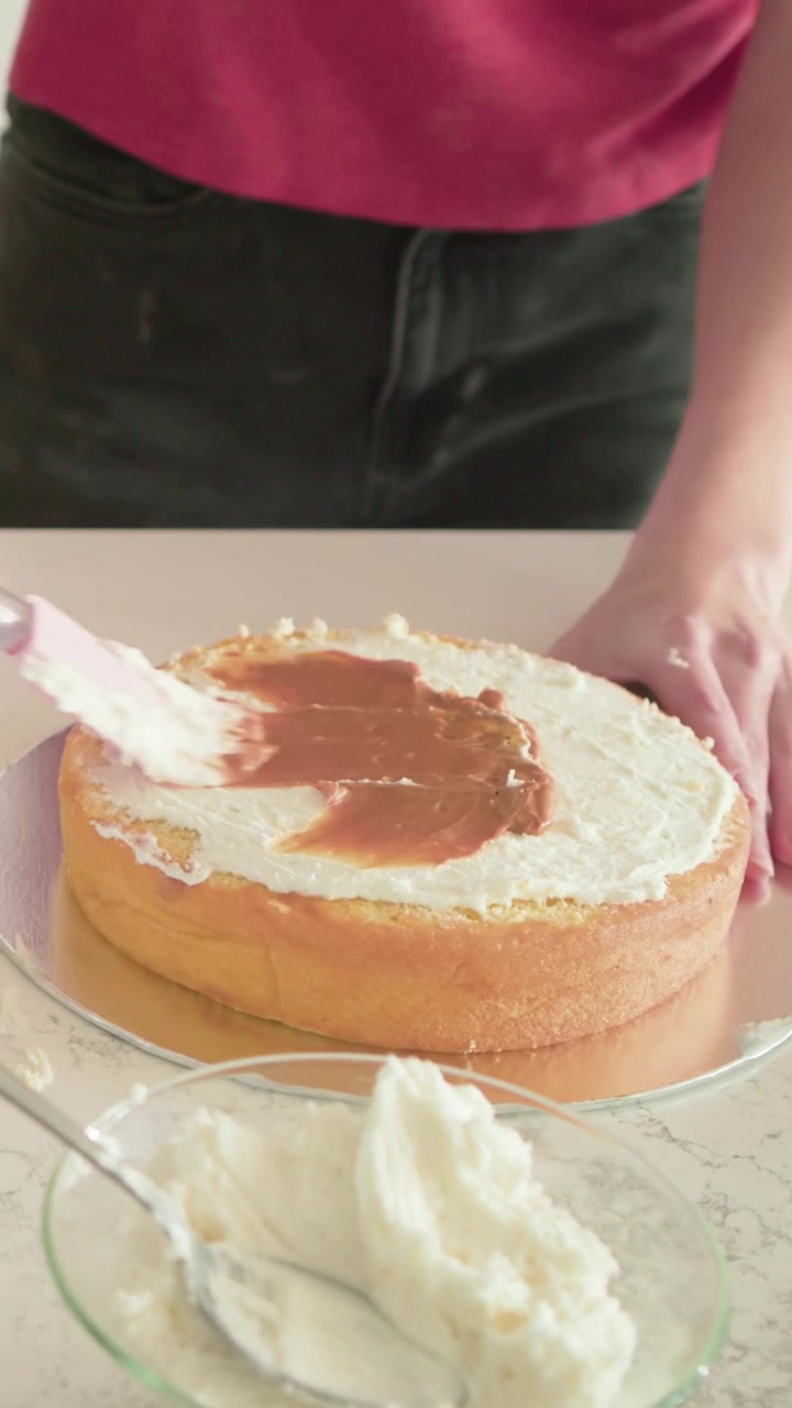Video verticale: Pasticcera con top rosa che prende delle bacche e decora una torta a casa con una spatola.