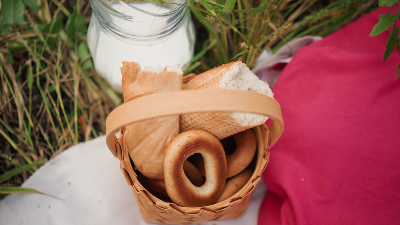 Primo piano della mano di una donna che si protende in un cesto intrecciato pieno di pane e bagel, accanto a un barattolo di vetro. La scena include erba, piante, tessuto bianco e una donna che indossa abiti rosa. Luce naturale soffusa.