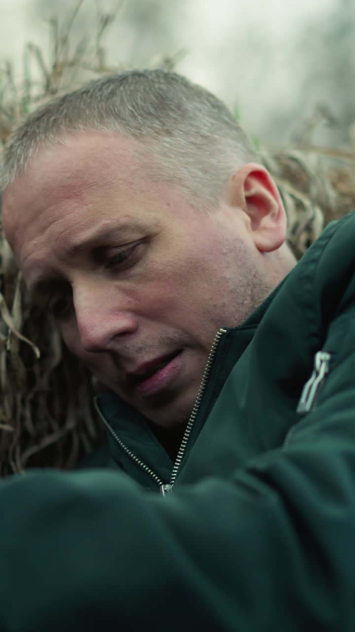 una vista cercana de un hombre con una chaqueta verde acostado en una zanja contra la hierba seca, sosteniendo una pistola con una expresión tensa, y mirando a su alrededor