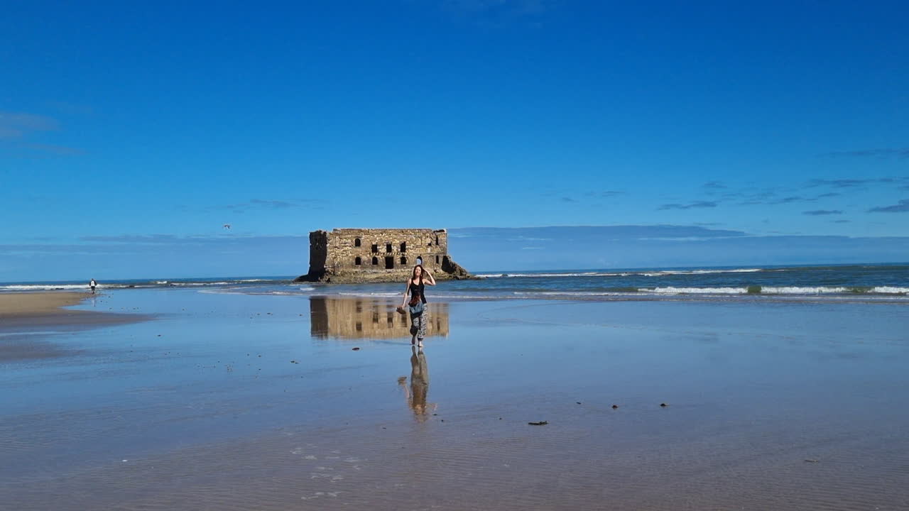 Tourist walking on wet sand of Tarfaya beach with Casa del Mar reflected in water