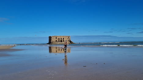 tourist walking on wet sand of tarfaya beach with casa del mar reflected in water