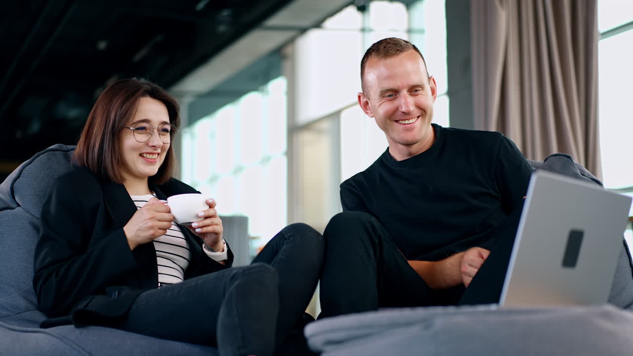 Positive Caucasian people sitting comfortably in bean bag chairs looking at laptop. Man and woman look at screen laughing. Low angle view.