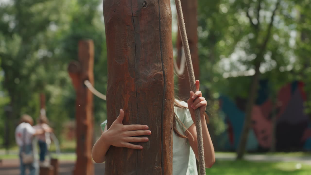 Figlia che si stabilizza su un palo di legno dopo una traversata con la corda, mani che abbracciano il palo e capelli intrecciati che cadono, momento di respiro e tranquilla resilienza nel parco, figure familiari in lontananza e calda luce estiva.