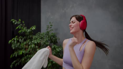 Close-up-of-a-happy-and-joyful-athletic-brunette-girl-wearing-a-purple-top-and-red-wireless-headphones-dancing-and-having-fun-while-listening-to-music-after-playing-sports-in-a-modern-apartment