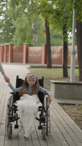 little girl with cerebral palsy sits in wheelchair holding hand of caring mother. young woman enjoys walking with daughter on road in city park