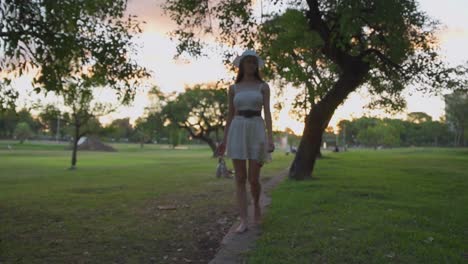 Beautiful-young-woman-in-hat-walking-barefoot-through-park-carrying-shoes-and-rose-in-her-hands