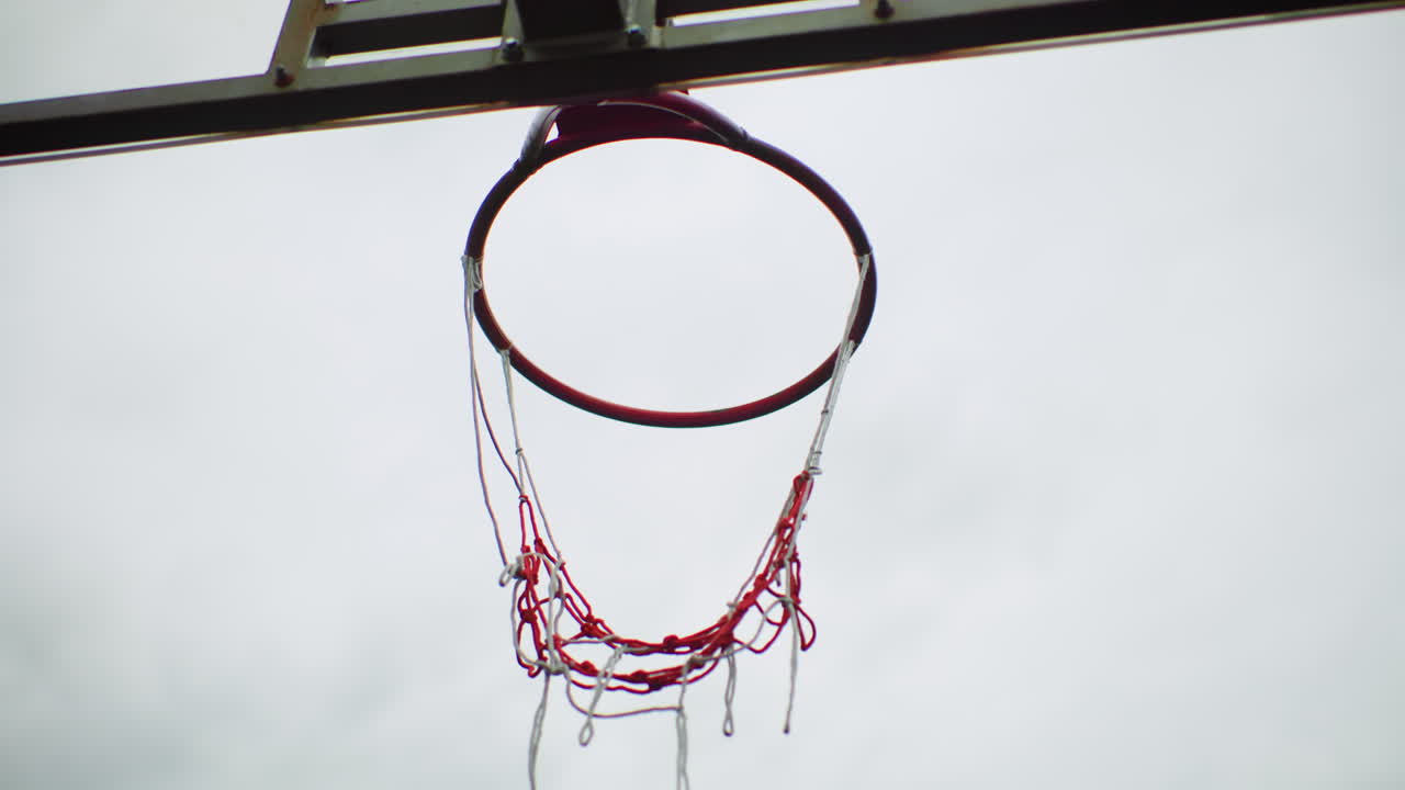 Cielo nuvoloso, canestro da basket all'aperto, sequenza di rumore di rete che mostra la palla che passa attraverso la rete a catena, impatto sul cerchio e rinculo della rete, sfondo di parco giochi urbano vuoto, toni attenuati e atmosfera nostalgica, rallentatore