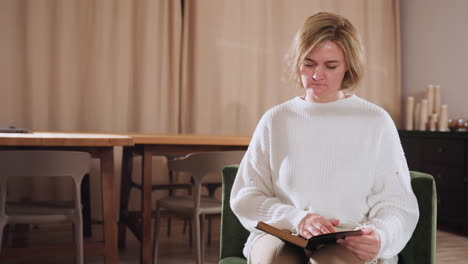emotional traveler sits on cushioned seat flipping through pages of book in warmly lit room with wooden tables and stacked paper cups in background, suggesting thoughtful moment of solitude