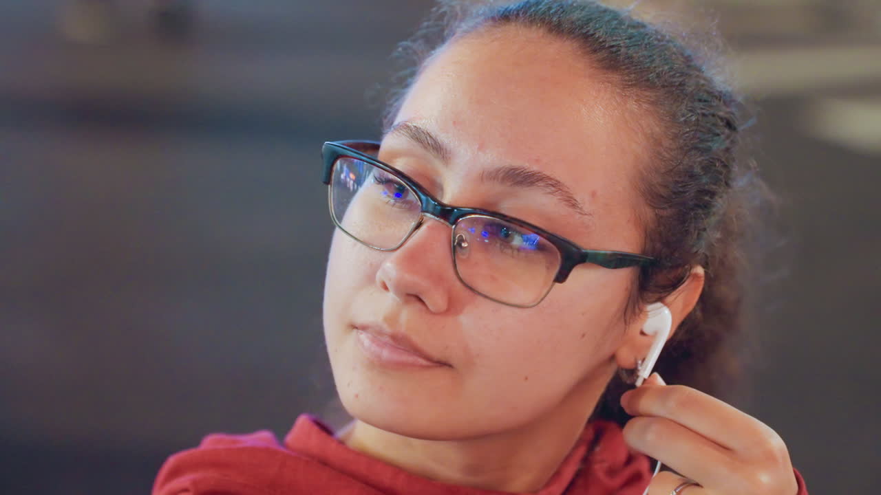 Young woman wearing glasses adjusting earphone in soft indoor light red hoodie closeup montage shows student inserting earbuds, commuter checking fit, podcast listener pausing to tune sound, attentive