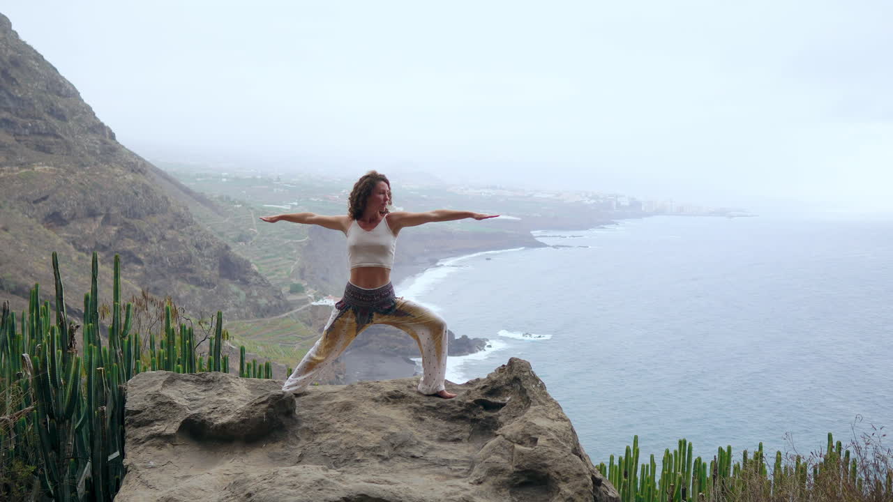 Amidst ocean, beach, and rocky mountains, a woman meditates in yoga's warrior pose, exemplifying motivation and an inspirational approach to fitness