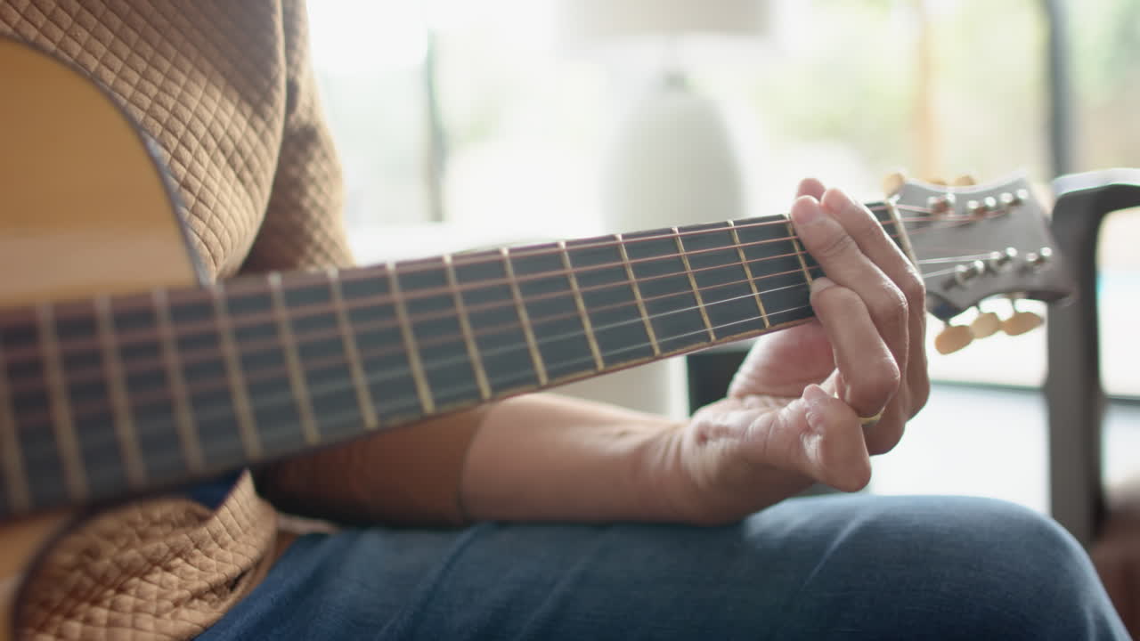sección media de un anciano biracial tocando la guitarra en una habitación soleada en casa, en cámara lenta