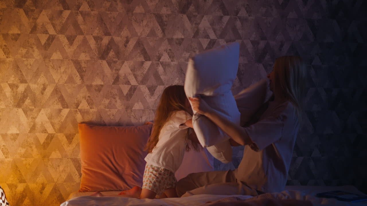 Child girl and mother in pajamas have pillow fight on bed