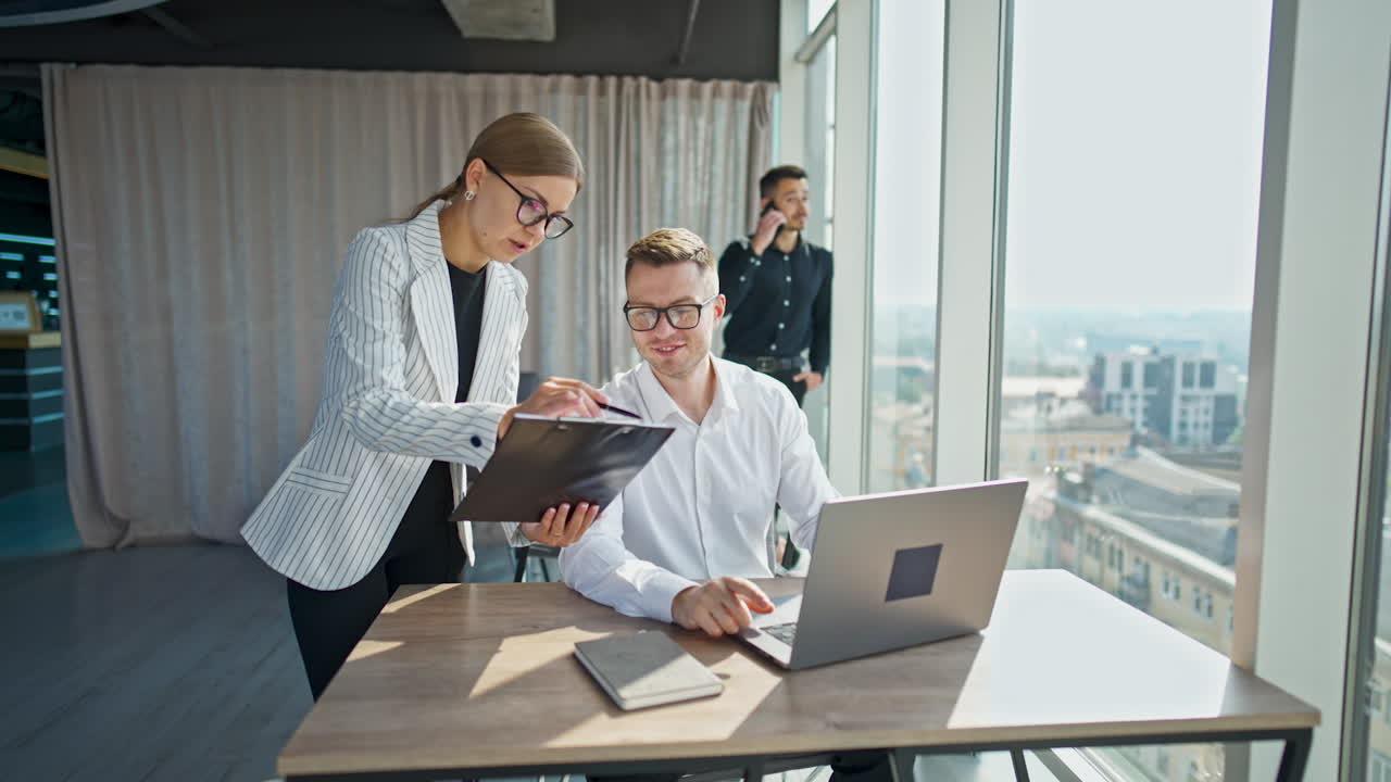 Female employee holding documents showing them to a male colleague. Man in black shirt stands near window at backdrop speaking on the phone.