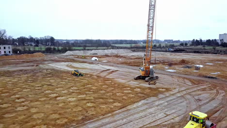 dynamic soil compaction - a tall crane drops a heavy weight on the foundation of a huge construction site - aerial pull back view