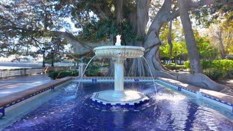 Una-Tranquila-Fuente-Centrada-En-Una-Piscina-De-Azulejos-Azules-Con-Agua-Cayendo-Suavemente-En-Cascada-Por-Sus-Niveles,-Con-El-Telón-De-Fondo-De-Un-Viejo-árbol-Majestuoso-Y-Un-Cielo-Despejado-En-Cádiz,-España.