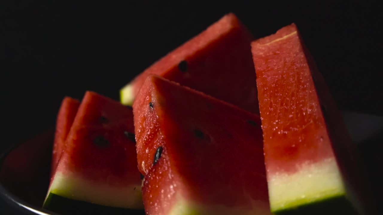 Close up zooming in video of gorgeous red colored and tasty delicious watermelon slices in triangle shaped with the green shell still on them while being put in front of a black studi background. Seed
