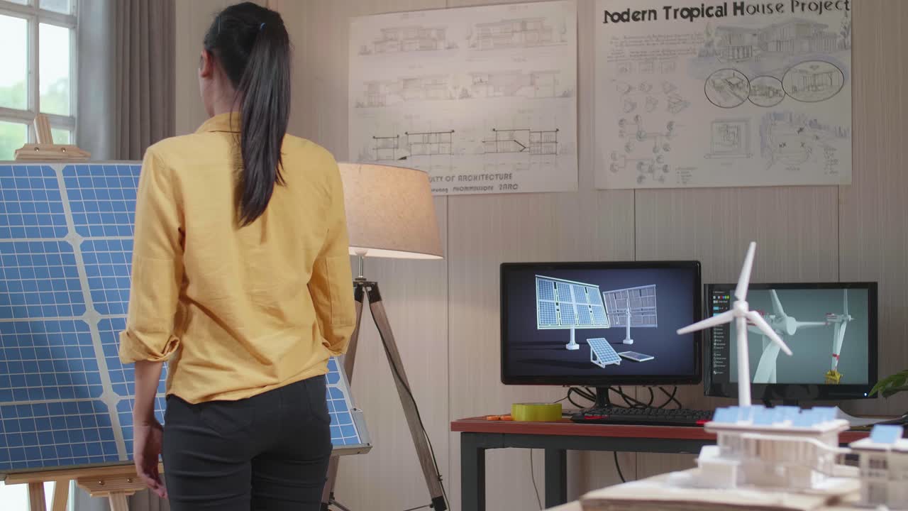 한 여성이 태양 패널과 풍력 터빈을 가진 모형 작은 집을 가진 방에서 태양 전지를보고 들어갑니다.