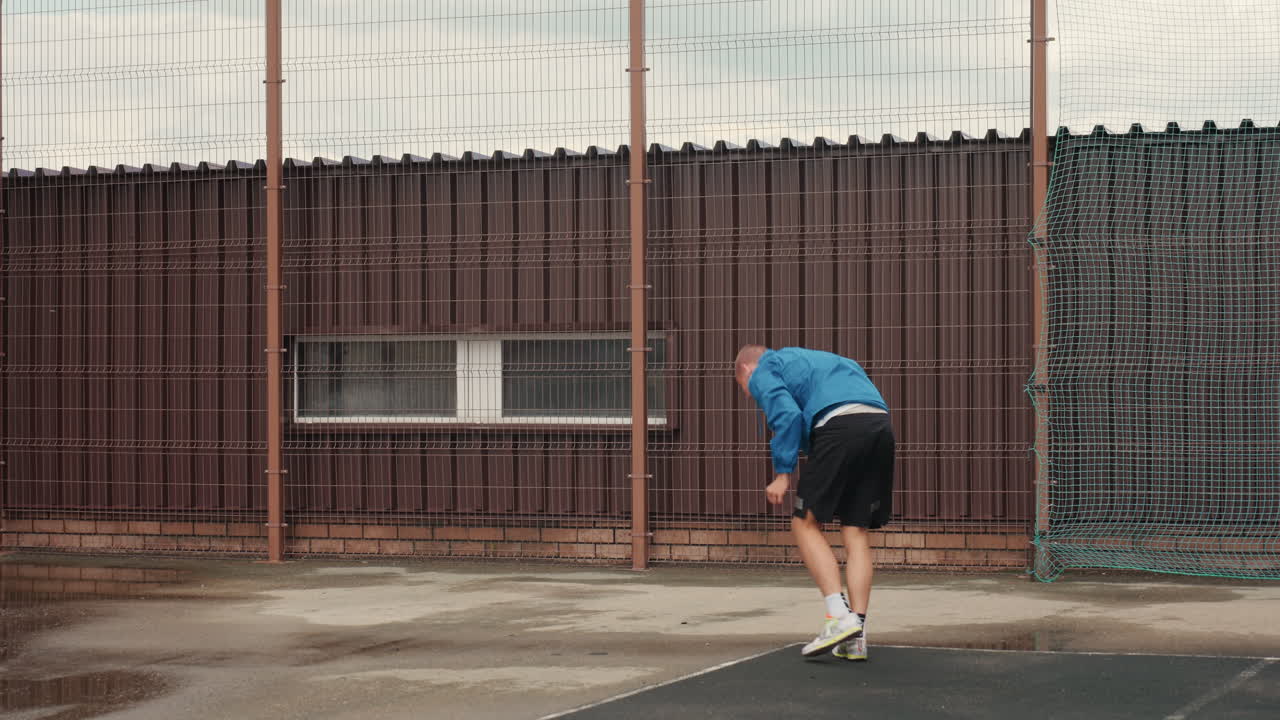 football player tying sneaker by fence, wet pavement and puddles under overcast sky, blue jacket and black shorts, solitary warmup bend followed by stand and walk toward rusty gate, raindrops