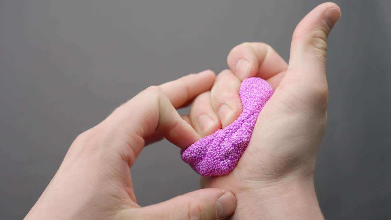 Hands squish and reshape lilac floam slime from a compact cube into an undefined mass, capturing sensory play and deformation on gray background, ASMR session, real time, static camera, close-up shot