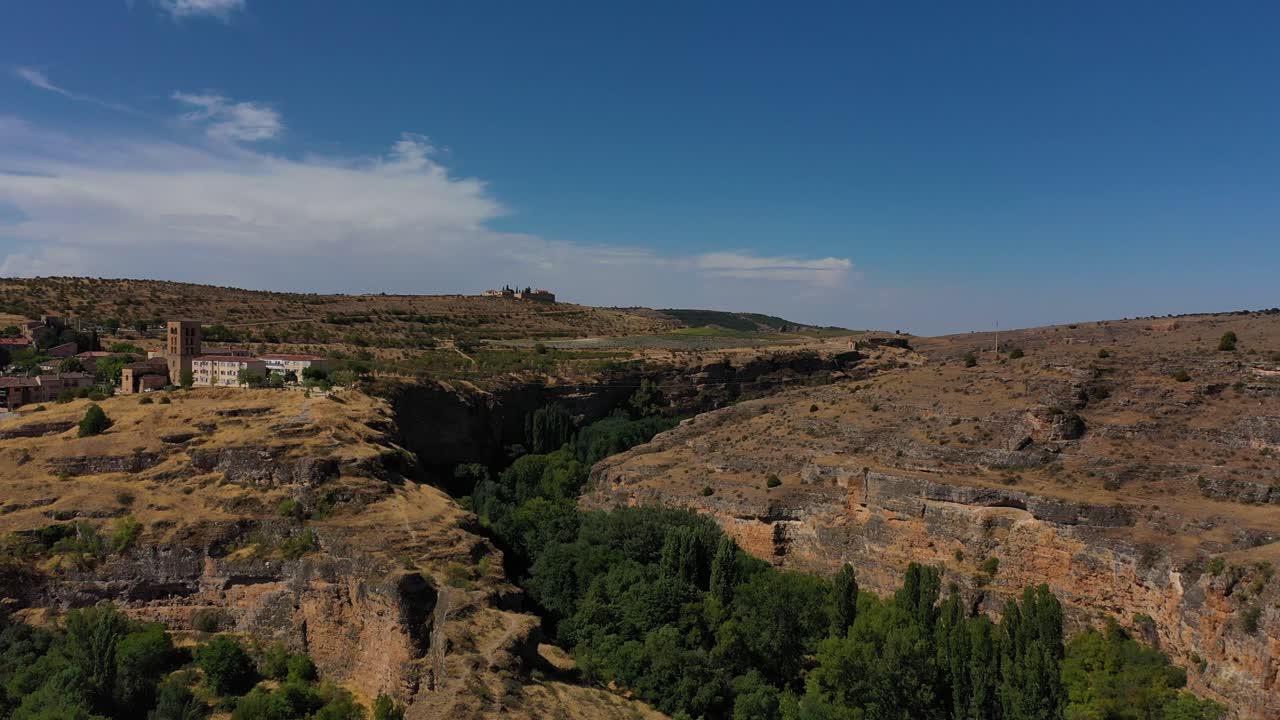 vuelo hacia atrás - ascenso a la vez sobre un cañón de garganta de piedra caliza con árboles en el curso del cañón y visualizando una ciudad con una torre en una mañana de verano con cielo azul segovia españa