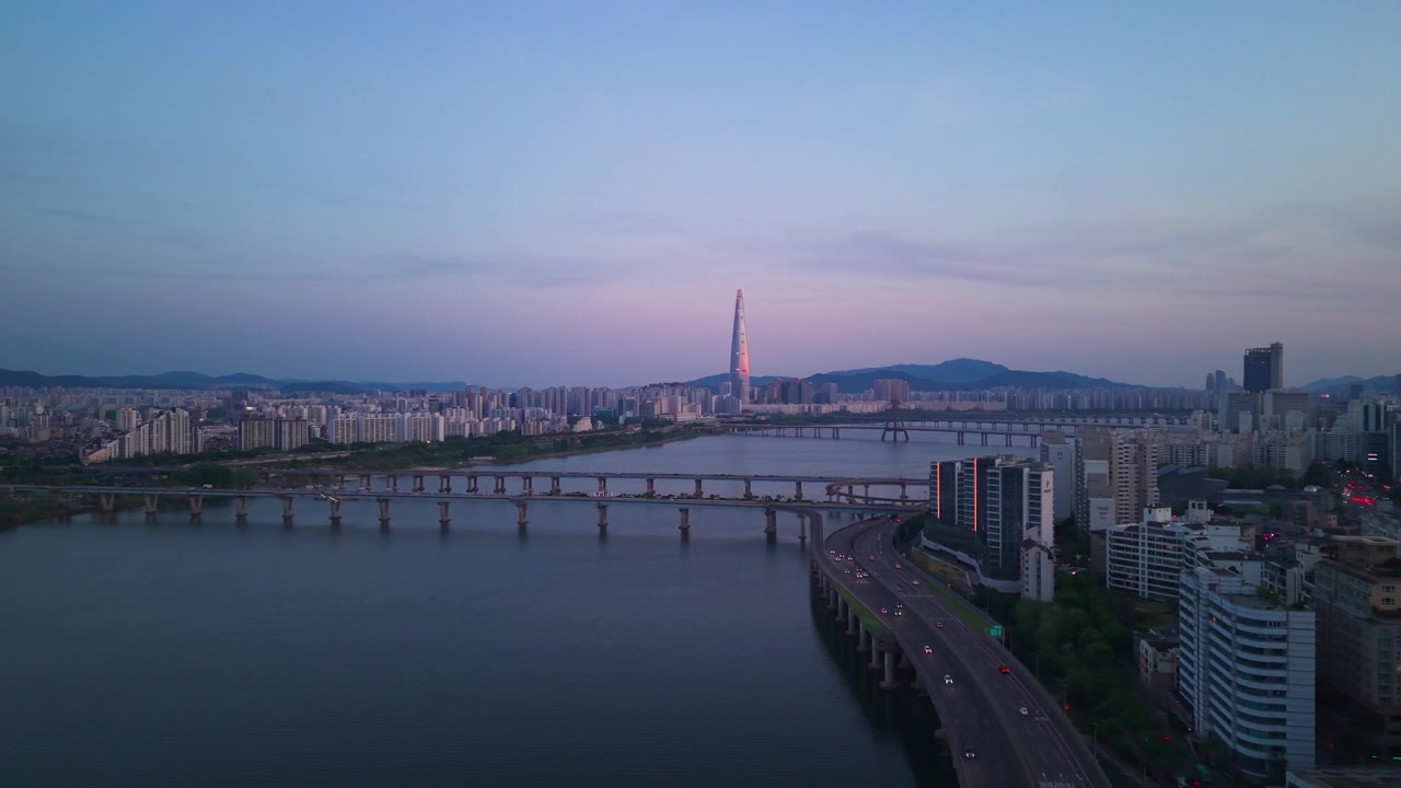 Panoramica aerea in prospettiva sulla sinuosa superstrada Gangbyeonbuk-ro al crepuscolo, che abbraccia i ponti del fiume Han, con la Lotte World Tower illuminata che si staglia contro lo skyline rosa-violaceo di Seul.