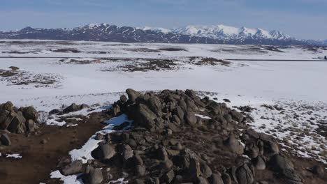 Drones-Volando-Sobre-Un-Campo-árido-Y-Lleno-De-Nieve,-Afloramientos-Rocosos-Con-Montañas-De-Dientes-De-Sierra-En-La-Distancia