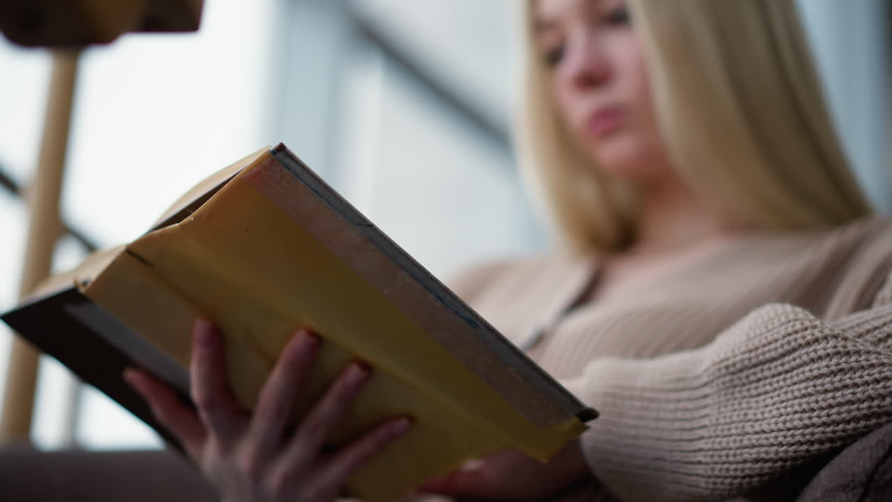 primer plano de una joven leyendo un libro mientras pasa a la siguiente página, sentada en un entorno acogedor, centrarse en las manos sosteniendo un libro desgastado, expresión pensativa borrosa