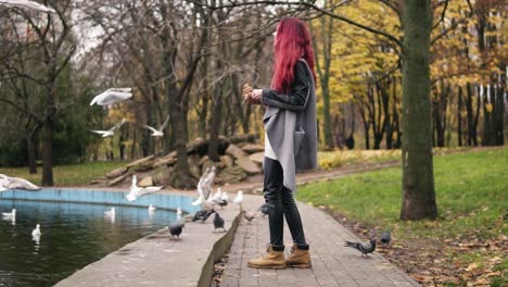 side view of red haired woman in warm coat feeding the gulls by the pond in park in autumn. slowmotion shot