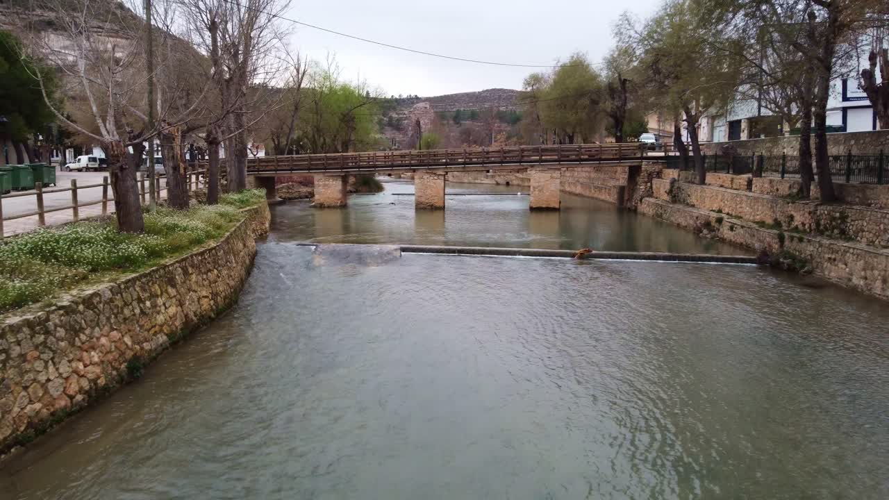 Imágenes de dron 4K volando bajo sobre el río Júcar en el pueblo de La Recueja. Muestra un puente peatonal, orillas de piedra y árboles ribereños en Castilla-La Mancha, España.