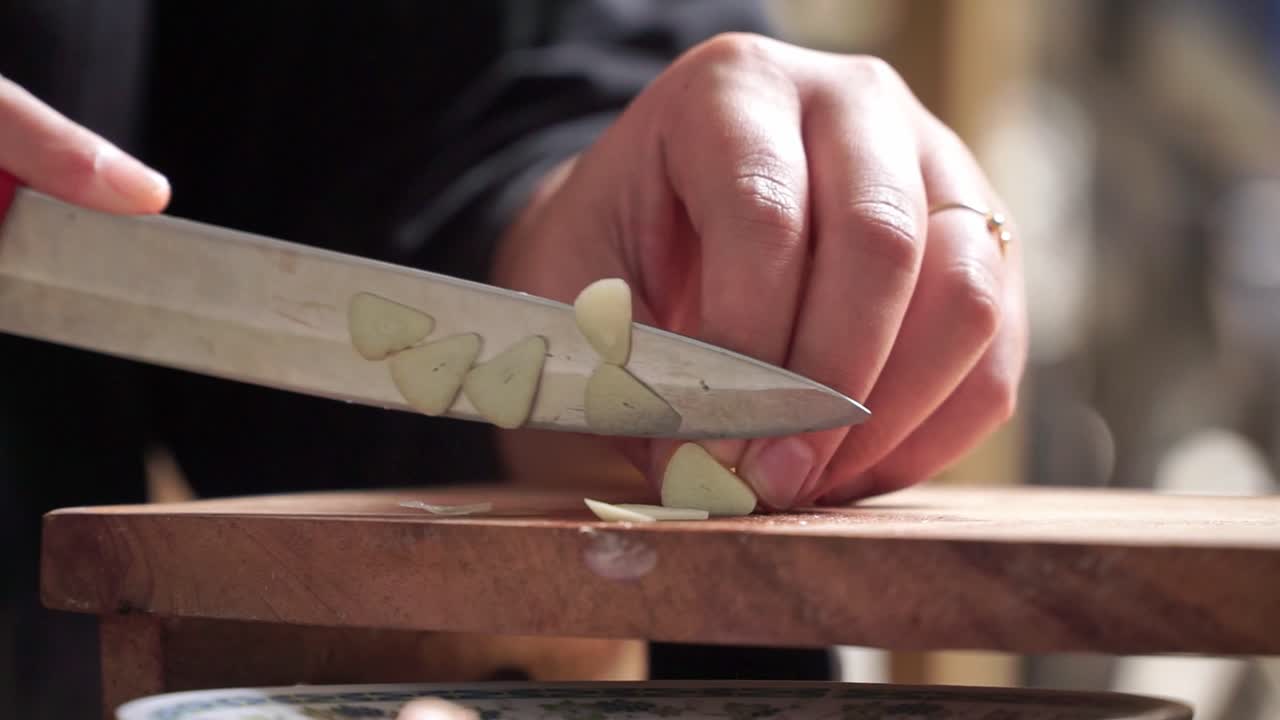 Close-up shot of hands slicing garlic on a wooden cutting board with a kitchen knife. Natural lighting and shallow depth of field emphasize a calm cooking moment.