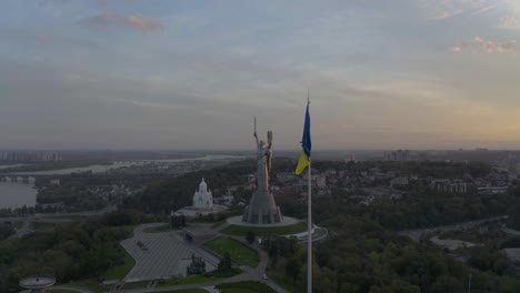 ukrainian flag fluttering in the wind with motherland monumental statue and temple of peace in the background at sunrise in kyiv, ukraine