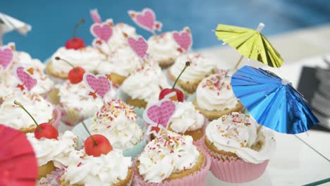 a bunch of colorful cupcakes with shinny umbrellas and sprinkles outdoors prepared for party with the swimming pool in the background