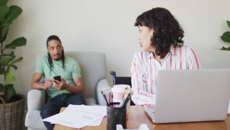 mujer biracial feliz en silla de ruedas usando computadora portátil y pareja masculina usando teléfono inteligente en la sala de estar