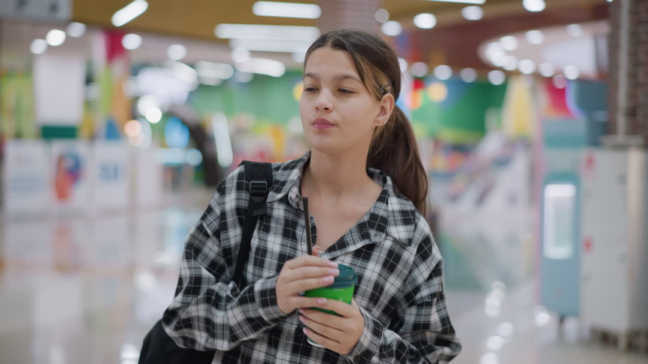 Young girl in checkered shirt sips juice as she walks through shopping mall, she carries backpack, gazing at bright festive lights in decorated modern retail space filled with colors and reflections