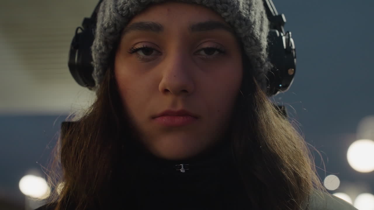 Nærbillede af ung kvinde med hovedtelefoner i aftenlys, blød bokeh-baggrund, eftertænksomt udtryk i vinterhue og jakke, filmisk stemningsfuld tone der formidler ensomhed og indadvendt lytning.