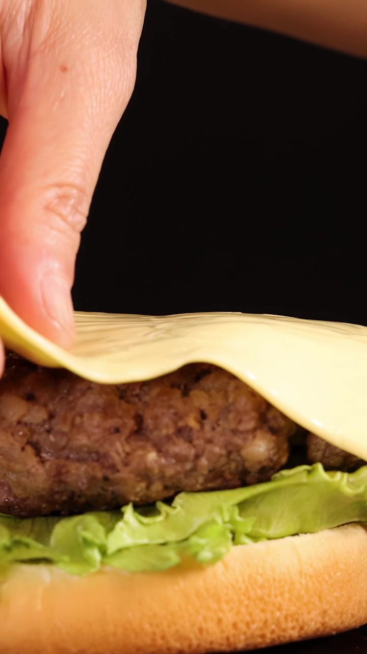 Una mano posiziona con cura una fetta di formaggio fuso su un hamburger di manzo cotto con lattuga su un panino da hamburger, sotto l'illuminazione da studio e uno sfondo nero.