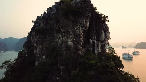 aerial shot moving up and over tall ha long bay island cliff to reveal boats on the water at sunset