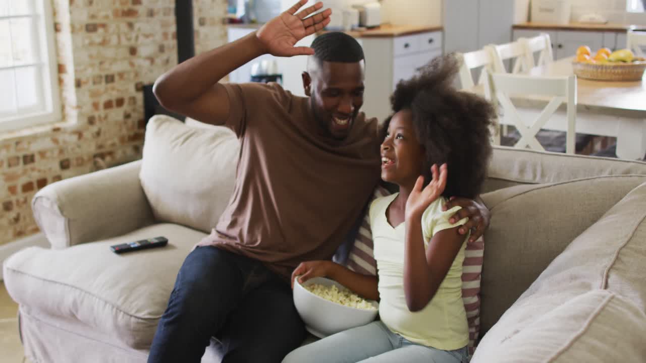 hija afroamericana y su padre en el sofá comiendo palomitas de maíz viendo televisión y dando cinco
