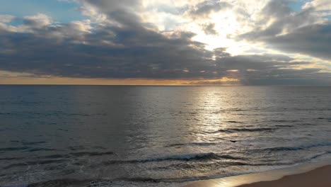 Wellen,-Die-Am-Ufer-Brechen,-Und-Ein-Sonnenuntergang-Am-Horizont-Hinter-Den-Wolken