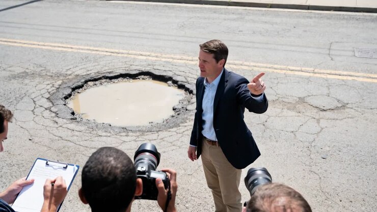 Raising finger, spokesman wearing blazer pointing to show pothole on road, with DSLRs and clipboard