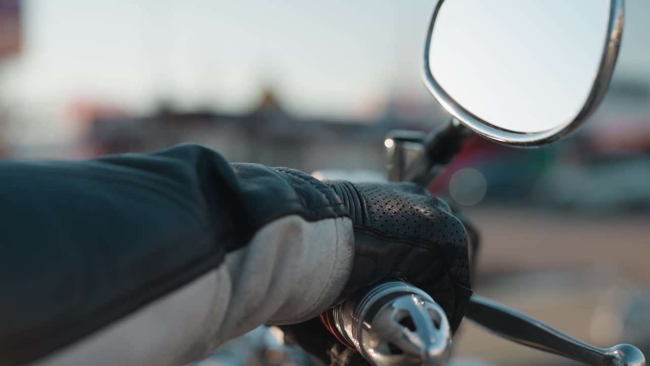 Close up of rider hand gripping motorcycle handle while wearing protective leather glove, mirror reflecting blurred city background, strong details emphasize control and lifestyle of motorcycling